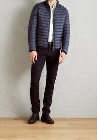 Marineblaue Steppjacke mit horizontalen Nähten, kombiniert mit schwarzer Jeans und braunen Schuhen. Die Jacke verfügt über einen Stehkragen und einen Reißverschluss.