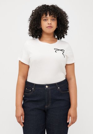 Een jonge vrouw met krullend haar draagt een witte t-shirt met een zwart strikontwerp, in combinatie met donkere jeans, staande tegen een lichte achtergrond.