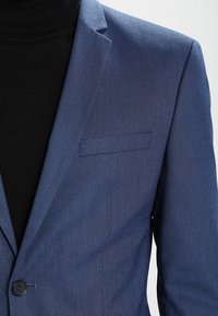 Blå texturerad kavaj med smal slag, en knappstängning och en bröstficka, bärs över en svart polotröja.
