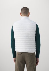 Hvid dunvest med kviltet design, stående krave og uden ærmer, båret over en mørkegrøn langærmet skjorte, parret med brune bukser.