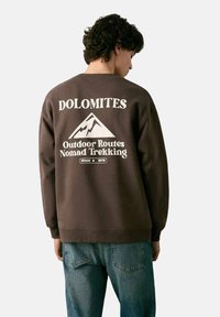 Bruine sweatshirt met witte grafische tekst en een illustratie van een berg op de achterkant. Geribbelde boorden en zoom. Gecombineerd met een blauwe spijkerbroek.