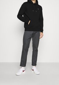 Zwarte hoodie met capuchon en voorzak; gecombineerd met grijze taps toelopende broek; witte sneakers met rode accenten; minimalistisch ontwerp.