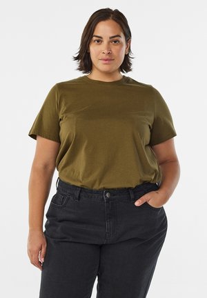 Femme aux cheveux courts et foncés portant un t-shirt vert olive rentré dans un jean noir taille haute, debout avec une main dans la poche.