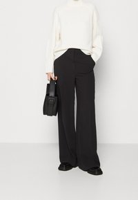 Svarta vida byxor med en mjuk textur, i kombination med en vit stickad tröja, och en liten svart handväska som hålls i höger hand.