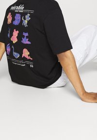 Musta t-paita, jossa on värikkäitä eläinkuvioita selkäpuolella, leikkisiä muotoiluja ja tekst banneri alla. Päällä valkoiset housut.