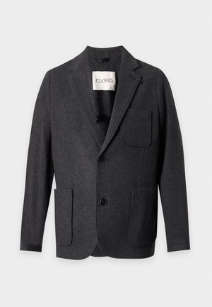 Homme portant un blazer noir, une chemise bleu marine foncé, un jean bleu clair et des mocassins en cuir noirs, debout avec une main dans la poche sur fond blanc.