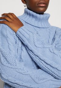 Maglione lavorato a maglia di un azzurro chiaro con colletto alto a costine, caratterizzato da motivi a treccia e diverse trame sulle maniche.