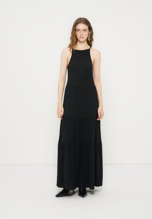 Jeune femme aux cheveux ondulés porte une robe maxi noire sans manches à col halter et des bottes noires à bout pointu, debout devant un fond blanc uni.