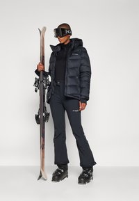 Sort pufferjakke og tætsiddende sorte bukser, udstyret med goggles og skiudstyr. Glat, isoleret stof med logoprægninger og strømlinet design.