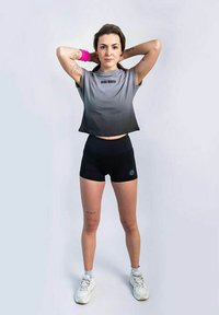 Crop top grigio sfumato con testo "BIDI BADU", abbinato a pantaloni corti neri a vita alta. Sneakers bianche e dettaglio del braccialetto rosa.