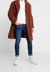 Abrigo largo marrón, texturizado, sobre un suéter blanco, combinado con jeans azules y zapatillas blancas. Presenta dos botones y un diseño de corte relajado.