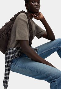 Brązowy plecak z falbanami, szary krótki sweter z dzianiny, nałożona niebiesko-czarna koszula w kratę oraz niebieskie dżinsy. Gładkie tekstury; swobodny krój.