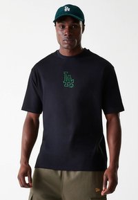 Svart bomulls-T-skjorte med en liten grønn LA-logo på brystet, kombinert med en mørkegrønn caps. Kortermet og med avslappet passform.