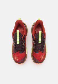 Rode en zwarte sportschoenen met een gaasstructuur, groene accenten en een bruine rubberen zool. Voorzien van vetersluiting en reflecterende details.