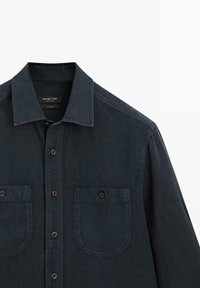 Mörkblå linne skjorta med en strukturerad krage, två bröstfickor, knäppning med knappar samt en subtil textur. Etikett synlig inuti kragen.