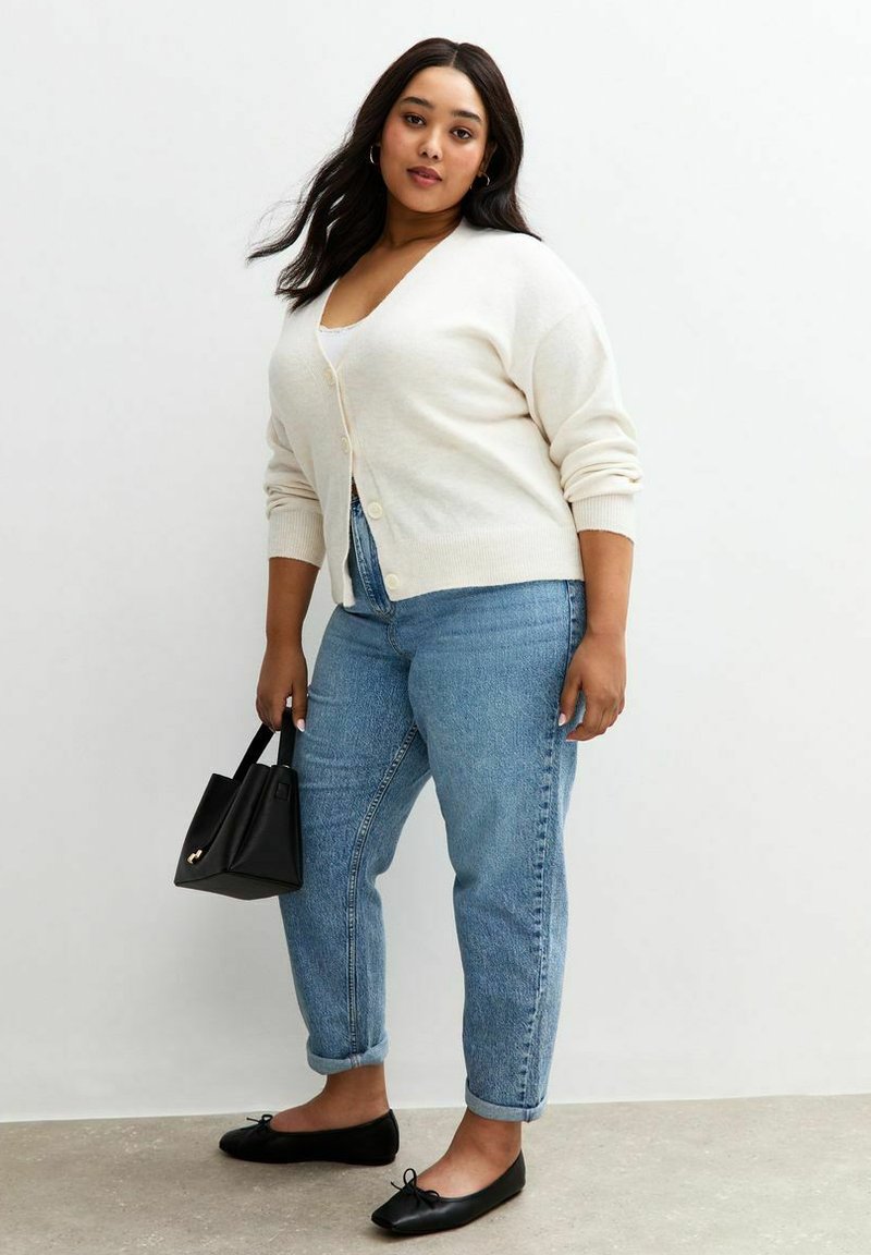 Cárdigan beige claro, abotonado, llevado sobre una blusa blanca, combinado con jeans de mezclilla azul claro. Unos zapatos planos negros y un pequeño bolso negro completan el conjunto.