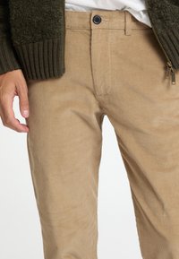 Καφέ κοτλέ παντελόνι με κατακόρυφες ραβδώσεις, διαθέτει μπροστινό κλείσιμο με κουμπί και πλαϊνές τσέπες. Το υλικό έχει απαλή, υφασμάτινη όψη.