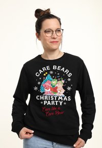 Felpa nera con un grafico colorato dei Care Bears a tema natalizio, fiocchi di neve e testo "Festa di Natale dei Care Bears." Materiale in cotone.