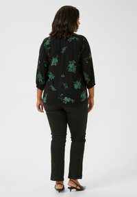 Schwarze Bluse mit grünem Floral-Muster, Dreiviertelärmel und lockerer Passform, kombiniert mit schwarzen Slim-Fit-Hosen und flachen Schuhen mit niedrigem Absatz.