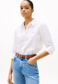 Camisa blanca de botones con cuello, que presenta un pequeño logo bordado. Combinada con jeans de mezclilla azules y un cinturón marrón, mostrando un look casual.