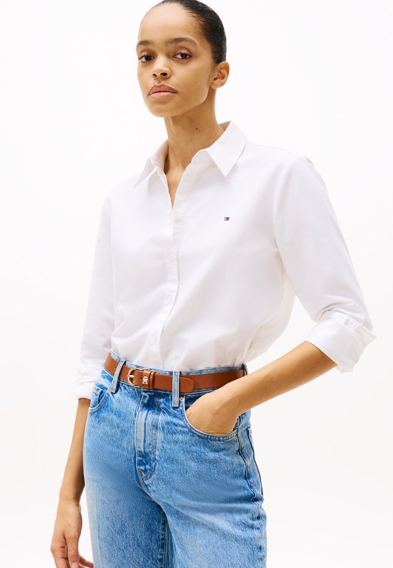 Camisa blanca de botones con cuello, que presenta un pequeño logo bordado. Combinada con jeans de mezclilla azules y un cinturón marrón, mostrando un look casual.