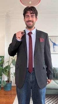 Mustabata musta blazeri maroon solmio ja taskuliina, sinivalkoraidallinen paita ja löysät farkut. Seisoo sisätilassa kasvin vieressä.