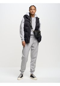 Moteris su pintais plaukais, vilkinti pilką džemperį su kapišonu, derančias sportines kelnes, juodą pūstinę liemenę, juodą per petį nešiojamą krepšį ir juodus sportinius batelius su baltais padais.