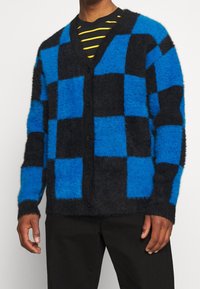 Sininen ja musta ruudullinen villatakki, pörröinen rakenne, nappikiinnitys, pitkät hihat, kontrastiväreillä musta ja sininen neliökuvio.