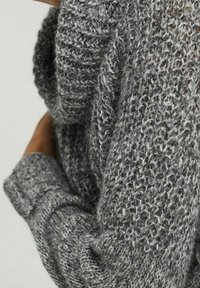 Pull en tricot gris avec une texture épaisse. Présente une coupe ample et des poignets côtelés, mettant en valeur un mélange de fibres gris foncé et clair.