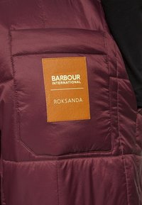 Burgunderfarbene gepolsterte Weste aus glattem Material mit einem braunen Etikett, das die eingeprägten Details "BARBOUR INTERNATIONAL" und "ROKSANDA" trägt.