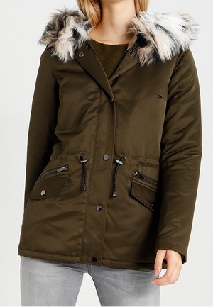 Olijfgroene parka met nepbontkraag, taillekoord, meerdere rits- en drukknoopzakken, gedragen over een donker sweater met lichtgrijze jeans.