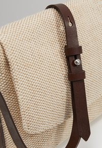 Beige texturerad tygväska med mörkbrun justerbar läderrem och silverfärgad metallknappdetalj på en enfärgad bakgrund.