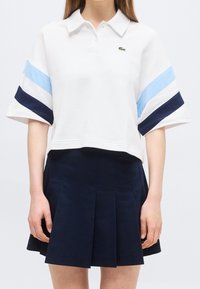 Polo court blanc avec des rayures bleu clair et bleu marine sur les manches et une jupe plissée bleu marine portée par une personne debout.