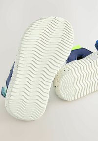 Sportschuhe mit einer strukturierten weißen Gummisohle, die gewellte Muster und schwarze Sprenkel aufweist; blaue und grüne Akzente im oberen Design.