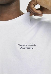 Hvid bomulds t-shirt med rund halsudskæring og korte ærmer, prydet med sort broderet tekst, der står "Naturens Kunstneriske Udtryk" på brystet.