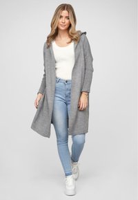 Grauer Strickcardigan mit Kapuze, gerippten Ärmeln und offenem Front, kombiniert mit einem weißen, eng anliegenden Oberteil und hellblauen Jeans. Weiße Sneakers.