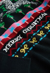 Mustalane kootud kampsun, millel on värvikas pühi muster, kus on zebra ja palmipuu kujutised. "HÄID JÕULE" valgete reljeefsete tähtedega.