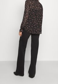 Kvinde iført sort blomsterprint transparent bluse og brede sorte bukser, stående med ryggen og siden mod en ensfarvet hvid baggrund.