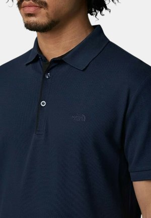 Uomo che indossa una polo blu navy con colletto e due bottoni, con un discreto logo ricamato The North Face sul petto a sinistra.