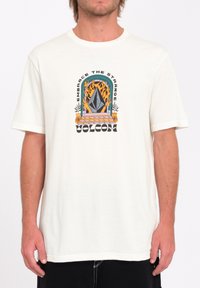 Camiseta de algodón blanca de manga corta con un diseño gráfico que presenta una forma de diamante abstracta, llamas y el texto: "Abraza lo Extraño Volcom."