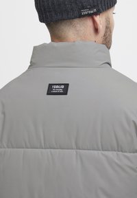 Grey puffer jacket with a high collar and a soft texture. Features a black label with white text on the back. Simple, functional design.
