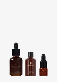 Trois bouteilles en verre ambré foncé avec des compte-gouttes et des bouchons noirs, étiquetées "Sérum Actif Chebula", "Essence Enzyme Microbiome" et "Huile Pure Radiance".