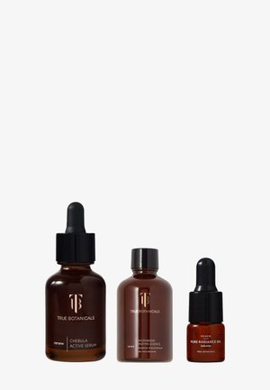 Drei dunkelbernsteinfarbene Flaschen mit schwarzen Tropfern und Kappen, beschriftet mit "Chebula Active Serum", "Microbiome Enzyme Essence" und "Pure Radiance Oil."