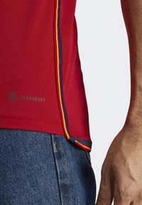 Červené sportovní tričko s texturou z síťoviny, s barevným bočním lemem (žlutá a modrá) a nenápadným logem "AEROREADY" u lemu.