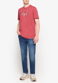 Rød bomulds t-shirt med grafisk print på forsiden, parret med blå denimbukser og sneakers i neutrale farver.
