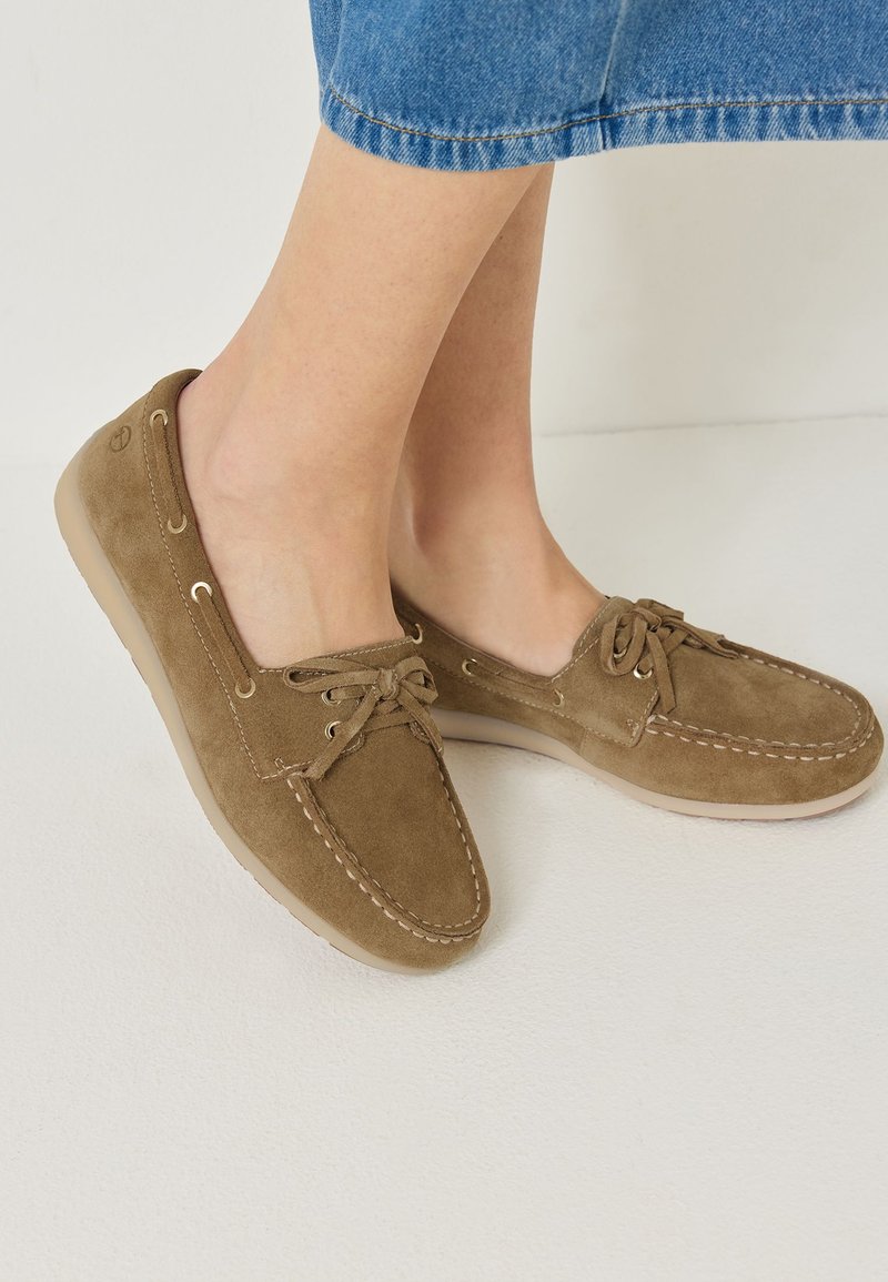 Pieds portant des mocassins en daim marron clair avec lacets, associés à un pantalon en jean bleu raccourci sur fond blanc.