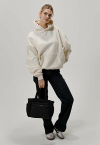 Sudadera de gran tamaño color crema con un bolsillo frontal, combinada con jeans oscuros y zapatillas de deporte grises. Bolso tote negro con un asa corta.