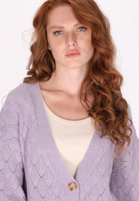 Cardigan lavorato a maglia lavanda con motivo a diamante, design a V, e un singolo bottone in legno. Indossato sopra una maglietta beige chiaro.