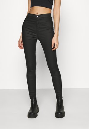 Vrouw draagt een zwarte skinny jeans met hoge taille, een zwarte crop top en zwarte enkellaarzen, staand tegen een effen witte achtergrond.
