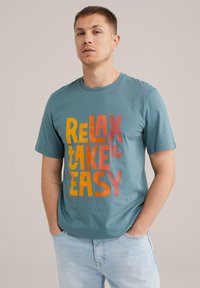 Türkiz pamut póló, amelyen merész, színes felirat található: "RELAX TAKE it EASY." Kerek nyakkal és rövid ujjakkal rendelkezik, lazább szabásban.